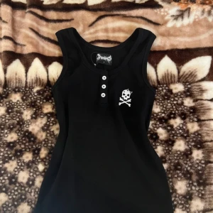 Svart ärmlös linnetopp med dödskalle - Svart deadstock vintage RARE ärmlös linnetopp med knappdetaljer framtill och vit dödskallebrodyr på bröstet. Perfekt för dig som gillar en edgy stil och vill sticka ut lite extra. Även på min vinted yuutabox!