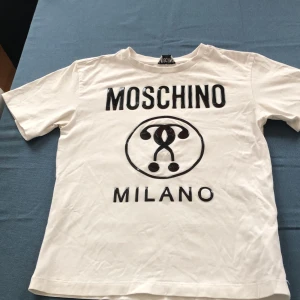 Vit t-shirt från Moschino Kid - Vit t-shirt från Moschino Kid med svart tryck framtill där det står 'MOSCHINO MILANO' och en stor logga i mitten. Klassisk rund halsringning och korta ärmar. Perfekt för dig som gillar statement-plagg och vill sticka ut.