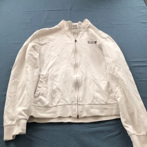Vit ziptröja från Emporio Armani EA7 - Vit ziptröja från Emporio Armani EA7 med hög krage och dragkedja framtill. Tröjan har två fickor på sidorna och logga på bröstet. Perfekt för en clean och sportig stil. Några fläckar på ena ärmen.