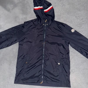 Moncler windbreaker  - Snygg marinblå vindjacka från Moncler med huva och dragkedja framtill. Jackan har en röd och vit rand på huvan och Moncler-logga på ärmen. Perfekt för blåsiga dagar och enkel att matcha med det mesta. Skick10/10. Storlek 2( motsvarar S-M) 