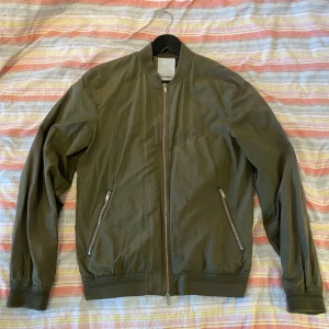 Olivgrön bomberjacka från J.Lindeberg - Säljer en stilren olivgrön bomberjacka från J.Lindeberg. Jackan har dragkedja framtill, två snedställda fickor med dragkedja och ribbade muddar vid ärmslut och nederkant. Perfekt för dig som gillar enkel och klassisk stil.