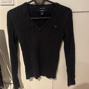 Svart kabelstickad tröja från GANT - Säljer en svart kabelstickad tröja från GANT i stretchig bomull. Tröjan har v-ringning, lång ärm och diskret GANT-logo broderad på bröstet. Perfekt till jeans eller kjol för en klassisk look.