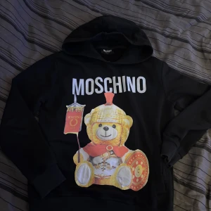 Moschino hoodie - Hoodien är använd och man ser det. Storleken på den är L fast det passar som en M. Jag är 183 och är lite kort på mig. Finns defekt som man kan se på bild 4 och 5. 