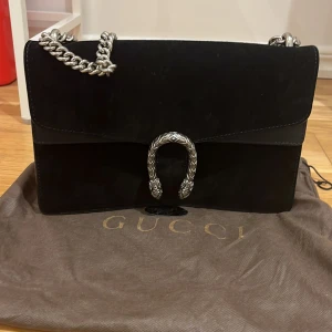 Svart axelväska med silverkedja - Säljer en svart axelväska med snygg silverkedja och dekorativt spänne framtill. Väskan har en stilren design och är tillverkad i skinn med mockadetaljer. Perfekt för dig som vill ha en lyxig accessoar. Inga tecken på användning 🖤