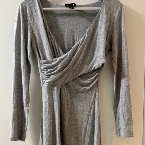 Grå omlottklänning med lång ärm - Säljer en stilren grå omlottklänning med lång ärm och djup v-ringning. Klänningen har en draperad omlottdesign framtill och är tillverkad i ett mjukt, stretchigt material som ger en smickrande passform.