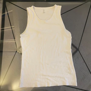 Vit ribbad linnetopp - Säljer en vit ribbad linnetopp i stretchigt material. Toppen är ärmlös och har en klassisk rund halsringning. Perfekt för varma dagar eller som lager under andra plagg.