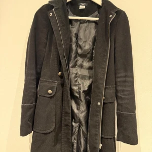 Svart kappa med knappar och dragkedja från Mouli - Säljer en svart kappa från Mouli med långa ärmar, stora fickor med dekorativa knappar och dragkedja framtill. Klassisk och stilren design med markerad midja och slits bak. Perfekt för kyligare dagar.