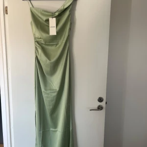 Adoore känning (ny med lappar) - Elegant, hellång klänning i en vacker ljusgrön nyans. Klänningen har en asymmetrisk halsringning med en draperad detalj som ger en sofistikerad touch. Den är designad för att framhäva figuren på ett smickrande sätt.  Oanvänd med alla lappar kvar. Nypris 1600kr