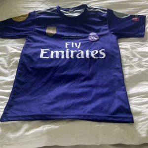 Real Madrid Ronaldo 7 fotbollströja - Säljer en blå Real Madrid fotbollströja med Ronaldo 7 på ryggen och Fly Emirates-tryck på framsidan. Tröjan har korta ärmar, klubbmärke och Champions League-detaljer. Perfekt för fans av Ronaldo och Real Madrid.
