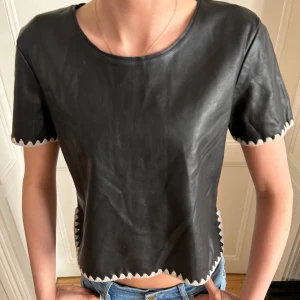 Svart skinnliknande topp med sicksack-detaljer - Cool svart topp i skinnliknande material med korta ärmar och sicksack-detaljer längs sömmarna. Croppad så slutar lite under naveln. Är från Zara och i storlek M