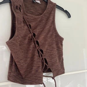 Brun croppad linnetopp med snörning - Säljer en brun croppad linnetopp med snörning framtill. Toppen har ett rutigt mönster och är ärmlös med rund halsringning. Perfekt för varma dagar eller lager på lager.
