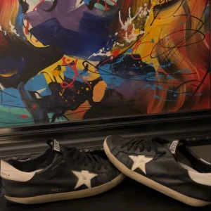 Svarta sneakers med vit stjärna från Golden Goose - Säljer ett par svarta sneakers från Golden Goose med vit stjärndetalj på sidan och vit häl. Skorna har snörning och platt sula, samt GGDB/SSTAR-tryck på sidan. Perfekta för en avslappnad och trendig stil.