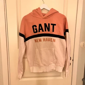 Rosa och vit hoodie från GANT - Säljer en snygg hoodie från GANT i rosa och vitt med svart detalj och texten 'GANT' och 'NEW HAVEN' framtill. Tröjan har huva och magficka, perfekt för en avslappnad stil.