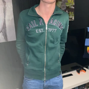 Grön zip-jacka från Sail Racing - Mörkgrön jacka med dragkedja från Sail Racing. Broderad logga och text på bröstet samt patch på ärmen. Två fickor med dragkedja och hög krage. Perfekt för en sportig och avslappnad stil. Han på bilden är 180 cm och väger 62 kg 