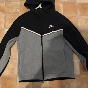 Nike grå och svart hoodie jacka - Nike jacka med huva i grått och svart, dragkedja framtill och två fickor. Vit Nike-logga på bröstet och vit detalj över bröstet. Sportig look med lång ärm och bekväm passform.