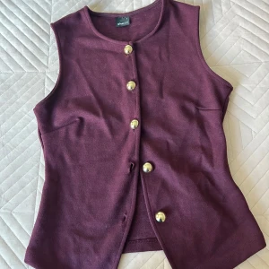 Vinröd väst med guldknappar - Vinröd väst från gina med fyra stora guldknappar framtill. Ärmlös och rund halsringning, i ett mjukt stickat material. Perfekt för att sommaren, helt i nya skick