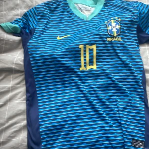 Blå Brasilien fotbollströja Neymar JR 10 - Snygg blå Brasilien fotbollströja med Neymar JR och nummer 10 i gult. Tröjan har korta ärmar, vågigt mönster i olika blå nyanser och Brasilien-emblem på bröstet. Perfekt för dig som älskar fotboll och vill sticka ut på planen.