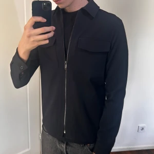 Overshirt  - Stilren svart/marinblå  jacka med dragkedja framtill och klassisk krage. Jackan har två bröstfickor med lock och långa ärmar med knapp vid ärmslut. Perfekt för en clean och enkel look. Storlek M. Det är en mörk marinblå färg 