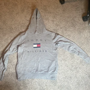 Grå hoodie från Tommy Hilfiger - Säljer en grå hoodie från Tommy Hilfiger med klassisk logga och text på bröstet. Tröjan har huva och ribbade muddar vid ärmslut och nederkant. Perfekt för en avslappnad och stilren look.