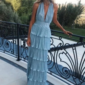 Ljusblå långklänning med volanger - Säljer en ljusblå långklänning med djup v-ringning och bred midja. Klänningen har flera lager volanger och är ärmlös med halterneck. Perfekt för bal eller fest! Materialet har en lätt glansig finish. 