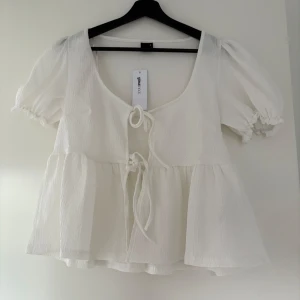 Vit blus med knyt  - Superfin vit blus från Gina Tricot med puffärmar och två knyt framtill. Blusen har en luftig och flowy passform, lätt strukturerat tyg och rundad halsringning. Perfekt för varma dagar och enkel att styla. Oanvänd med prislapp 