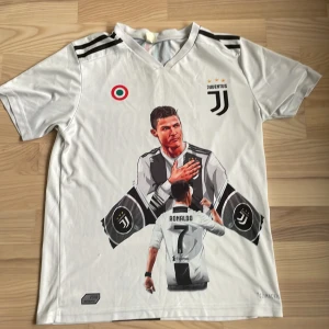 Juventus fotbollströja med Ronaldo-tryck - Vit kortärmad Juventus fotbollströja med stort tryck av Ronaldo på framsidan. Tröjan har svarta detaljer på axlarna och Juventus-logga på bröstet. Perfekt för fans av Ronaldo och Juventus.