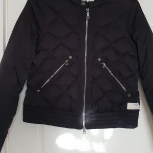 Svart quiltad pufferjacka från Reason - Säljer en svart quiltad pufferjacka från oddmolly med två snedställda fickor med dragkedja och silverfärgad dragkedja framtill. Jackan har lång ärm och kortare passform, perfekt för kyliga dagar. 0/xs