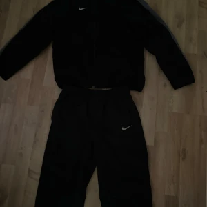 Svart träningsjacka från Nike - Svart träningsjacka från Nike med dragkedja och gråa detaljer längs ärmarna. Klassisk sportig look med Nike-logga på bröstet. Perfekt för träning eller chill. Jackan har lång ärm och normal passform.