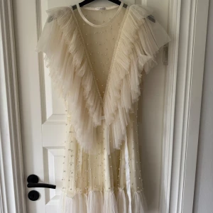 Vit volangklänning med pärlor från Ida Sjöstedt - Superfin vit klänning från Ida Sjöstedt med v-ringning och stora volanger framtill och baktill. Klänningen är dekorerad med små pärlor och har en lätt transparent känsla med tyll och omlottkänsla. Perfekt för speciella tillfällen!