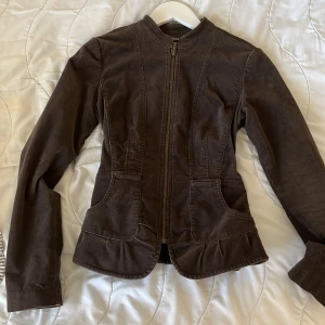 Brun bikerjacka från Mexx - Säljer en brun vintage bikerjacka från Mexx i manchester med dragkedja framtill, volanger längst ner och knappdetalj bak. Jackan har lång ärm, markerad midja och två fickor framtill. 