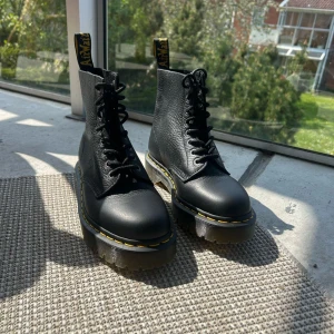 Svarta boots från Dr. Martens - Klassiska svarta boots från Dr. Martens med grov sula och gul söm runt sulkanten. Skorna har snörning framtill, rund tå och är tillverkade i skinn. Perfekta för dig som gillar en tuff och tidlös stil.