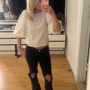 Vit blus med puffärmar och rosetter i ryggen - Säljer en vit blus med puffiga trekvartsärmar och två svarta rosetter i ryggen. Blusen är croppad och har en rak passform. Perfekt för dig som vill ha en trendig och söt topp med fina detaljer.