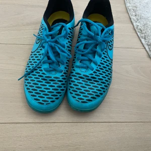 Blå Nike Magista fotbollsskor - Säljer ett par blå Nike Magista fotbollsskor med svart mönster och snörning. Skorna har låg profil, rund tå och vit sula. Perfekta för inomhusfotboll eller träning. Snygg design med svart Nike-logga på sidan.