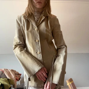Beige mockajacka med fransar - Mammas beige/gröna mockajacka med fransar. Är från USA från 90-talet. Den är tajt, men riktigt bekväm. Perfekt för dig som vill ha en western look! Storlek L, men passar nog S/M
