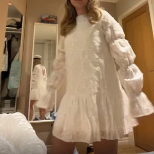 Vit volangklänning med öppen rygg - Superfin vit klänning med volangdetaljer och långärmade puffärmar. Klänningen har en öppen rygg med knytband upptill och är i kort modell. Perfekt för student eller sommarens festligheter!