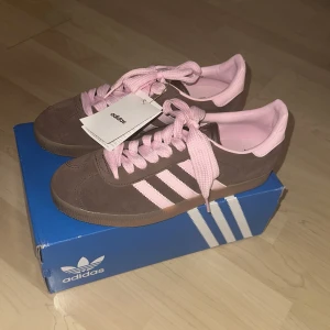 Adidas Gazelle brun och rosa sneakers - Säljer ett par Adidas Gazelle sneakers i brunt mocka med ljusrosa detaljer och snörning. Skorna har klassiska tre ränder på sidan och kommer med extra bruna och rosa skosnören. Skorna är i HELT nytt skick, och har inte används en ända gång. Skorna är även ganska små i sin storlek och går att ha i storlek 41-42