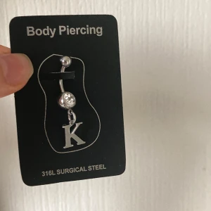 Navelpiercing med bokstav K och sten - Snygg navelpiercing i kirurgiskt stål med en glittrig sten och ett hängande K. Perfekt accessoar för dig som vill ha något personligt och stilrent.
