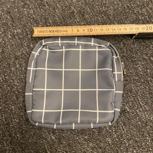 Grå-rutig liten necessär  - Säljer en liten grå necessär med vitt rutmönster. Perfekt för småprylar eller som accessoar. Necessären har dragkedja och är tillverkad i syntetmaterial. Enkel och stilren design.