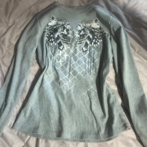 Grå ribbad långärmad tröja med döskalletryck - Säljer en grå ribbad långärmad tröja med stort tryck av två döskallar, fjädrar och kors på framsidan. Tröjan har ett rutigt mönster i bakgrunden. Perfekt för dig som gillar unika detaljer.