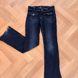 Mörkblå bootcut jeans med fickdetaljer - Mörkblå jeans med bootcut passform och snygga fickor framtill med lock och knappar. Jeansen har låg midja och klassiska bakfickor. Perfekta för en avslappnad och trendig look.