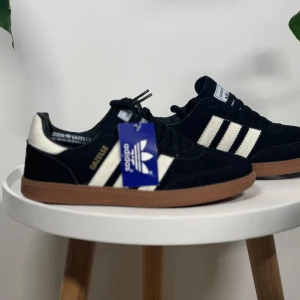 Adidas Gazelle sneakers svart/vit - Säljer ett par klassiska Adidas Gazelle sneakers i svart mocka med vita ränder och detaljer. Skorna har brun gummisula och tydlig Adidas-logga på tungan. Perfekta för dig som gillar retrostil och streetwear. Finns från storlek 40-45.