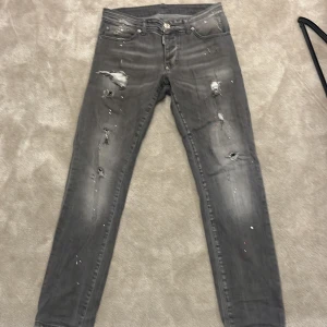 Grå jeans från Dsquared2 - Säljer ett par grå jeans från Dsquared2 med slitningar, hål och färgstänk. Byxorna har fem fickor, knappgylf och en röd DSQ2-detalj bak. Snygg, smal passform och coolt distressed utseende.