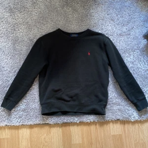 Svart sweatshirt från Polo Ralph Lauren - Klassisk svart sweatshirt från Polo Ralph Lauren med rund halsringning och röd broderad logga på bröstet. Tröjan har långa ärmar och ribbade muddar vid ärmslut och nederkant. Perfekt för en stilren och avslappnad look.