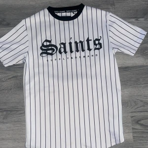 Vit randig t-shirt från Supply & Demand - Säljer en vit t-shirt med svarta vertikala ränder och texten 'Saints' i svart på bröstet. T-shirten har rund halsringning med svart kant och är kortärmad. Perfekt för en sportig och trendig stil.