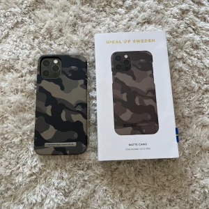  kamouflage skal från iDeal of Sweden - Snyggt mobilskal med matt camo-mönster i svart, grått och grönt från iDeal of Sweden. Passar iPhone 12/12 pro. Skalets material är hårdplast och har en metallisk logga längst ner. Perfekt för dig som vill ha en stilren och tålig look.