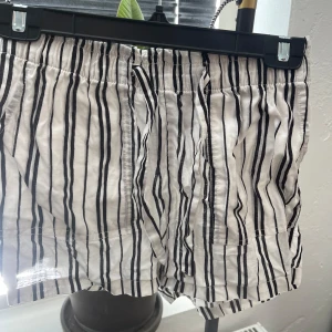 Randiga shorts från H&M - Säljer ett par vita shorts med svarta ränder från H&M. De har elastisk midja med snörning och är tillverkade i ett tunt, skönt bomullsmaterial. Perfekta för varma dagar och har en avslappnad passform.