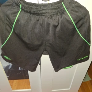 Svarta träningsshorts från Umbro med gröna detaljer - Svarta träningsshorts från Umbro med gröna kontrastsömmar och elastisk midja. Perfekta för sport eller gym, tillverkade i lätt och ventilerande syntetmaterial. Klassisk sportig look med Umbro-logga på benet.