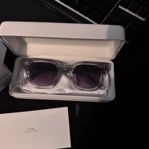 Transparenta solglasögon från Chimi - Snygga solglasögon från Chimi med transparent båge och mörka glas. Rektangulär form som ger en modern look. Kommer i ett vitt fodral.