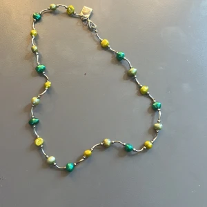 Halsband med gröna och gula pärlor - Handgjort halsband med oregelbundna pärlor i gröna och gula nyanser, separerade av små silverfärgade kulor och stavar. Halsbandet har ett diskret lås och en liten fyrkantig bricka vid låset.