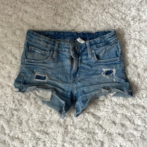 Blå jeansshorts från &Denim - Snygga blå jeansshorts från &Denim med slitna detaljer och fransig kant. Klassisk femficksmodell med låg midja och råa kanter. Perfekta för sommaren och en avslappnad stil.